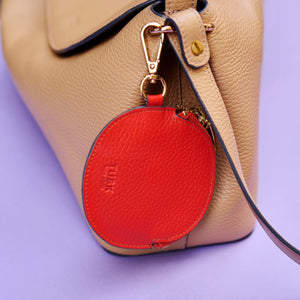 Orange leather round pouch charm attached to a beige handbag on a purple background - ORANGE