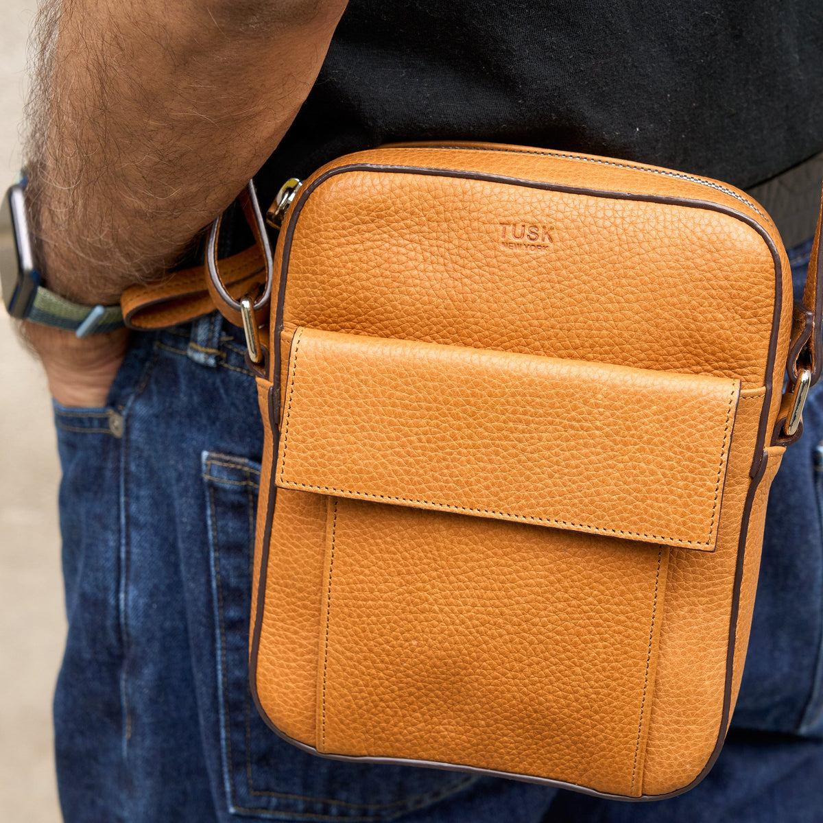 Brown leather crossbody bag with 'TUSK' branding worn by a person.