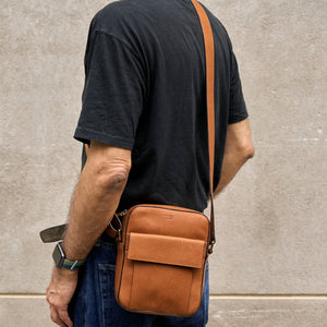A man viewed from behind wearing a black t-shirt and blue jeans, carrying a tan leather crossbody bag with a front pocket and a matching leather strap against a concrete wall - TOAST