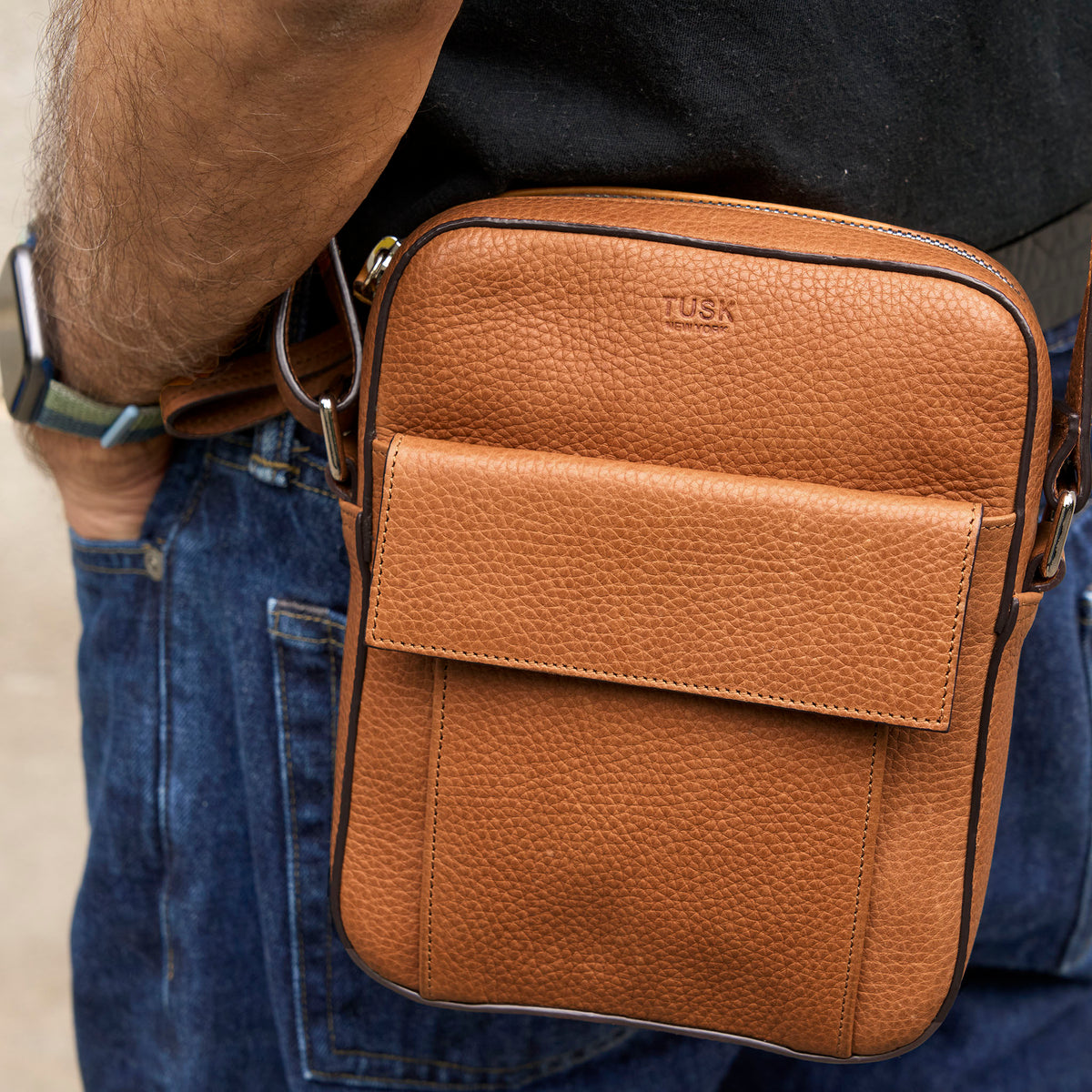 A tan pebbled leather crossbody bag with a front flap pocket, worn crossbody on a person's hip. The brand name 'TUSK' is subtly embossed on the upper right of the bag - TOAST