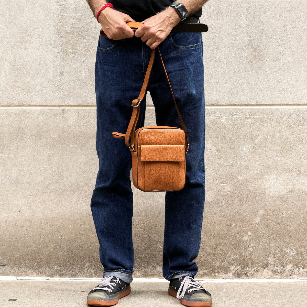 A person wearing dark blue jeans and black sneakers holds a tan leather crossbody bag against their hip, standing in front of a textured concrete wall - TOAST