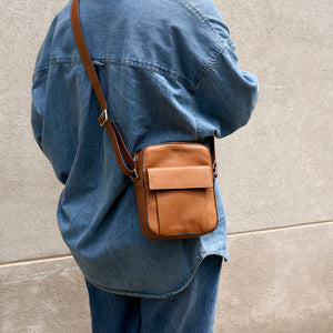 A person wearing an oversized blue denim shirt and jeans carries a small tan leather crossbody bag with a flap front and adjustable strap against a light concrete wall - TOAST