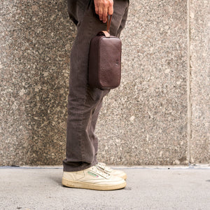 Person holding a brown toiletry bag against a textured wall - CHOCOLATE