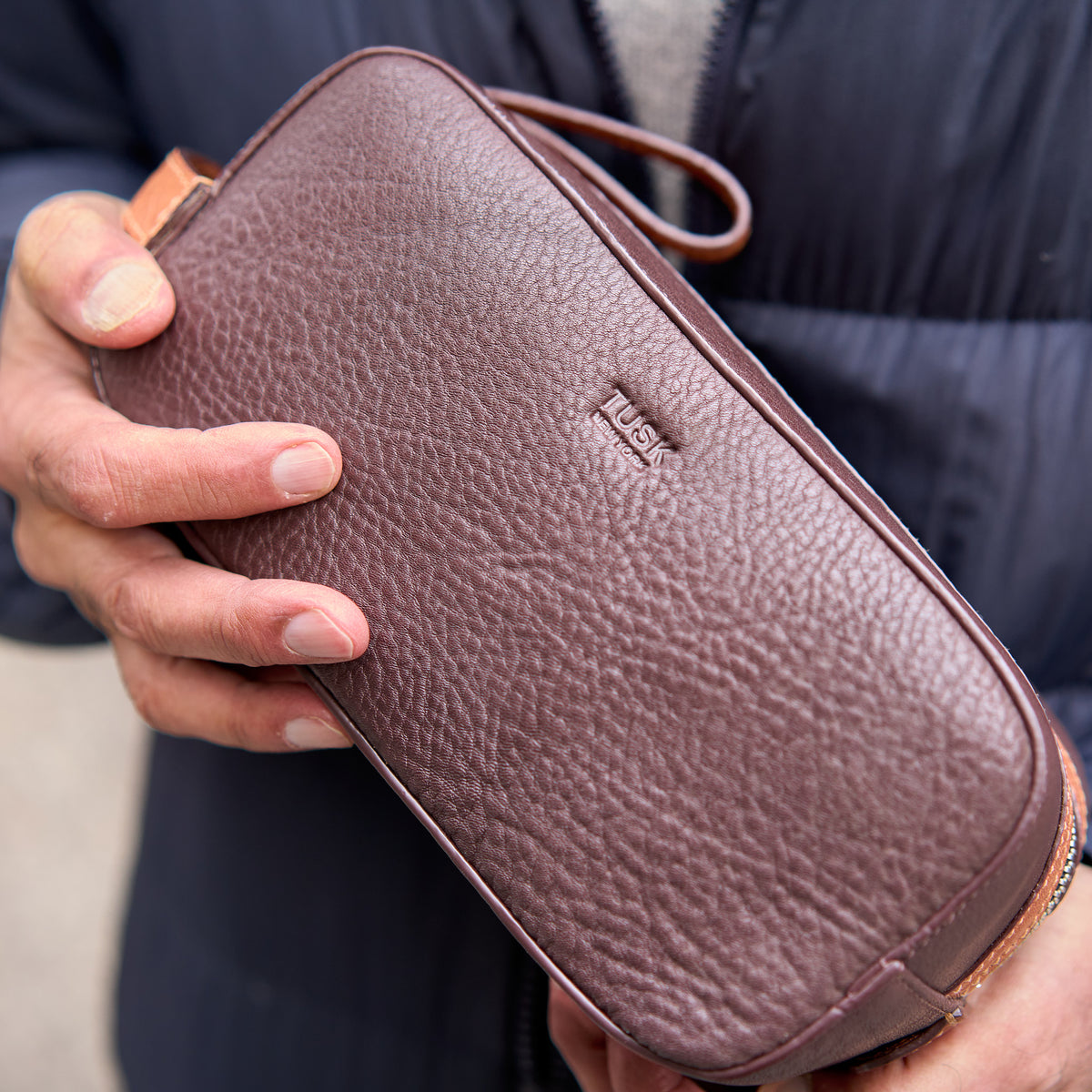 Brown leather toiletry bag held by a person with a blurred background - CHOCOLATE