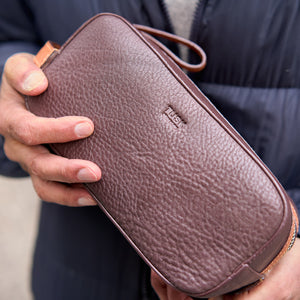Brown leather toiletry bag held by a person with a blurred background - CHOCOLATE