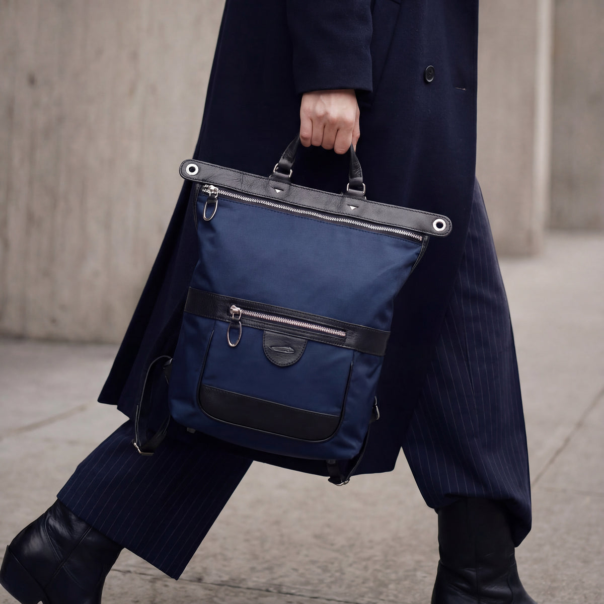 Close-up of a person walking outdoors in a dark navy pinstripe suit and black boots, carrying a structured navy blue nylon Tusk backpack by its black leather top handle. The backpack features black leather trim, a zippered front pocket, and silver hardware.- MIDNIGHT