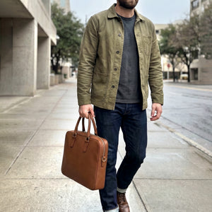 Man walking on a sidewalk holding a brown leather briefcase, wearing an olive green jacket and dark jeans - TOAST