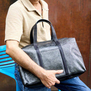 A man wearing a beige polo shirt and blue jeans holds a large, dark gray textured leather tote bag with black handles and trim, seated on a blue plastic chair against a rusty brown metal wall - SILVER