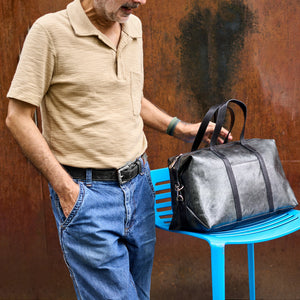 A man wearing a beige polo shirt and blue jeans stands beside a bright blue chair, holding a dark gray distressed leather duffel bag. The background is a textured rusted metal wall - SILVER