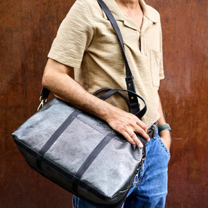 A person wearing a beige short-sleeve polo shirt and blue jeans carries a large gray leather duffel bag with black straps and trim against a textured brown wall - SILVER