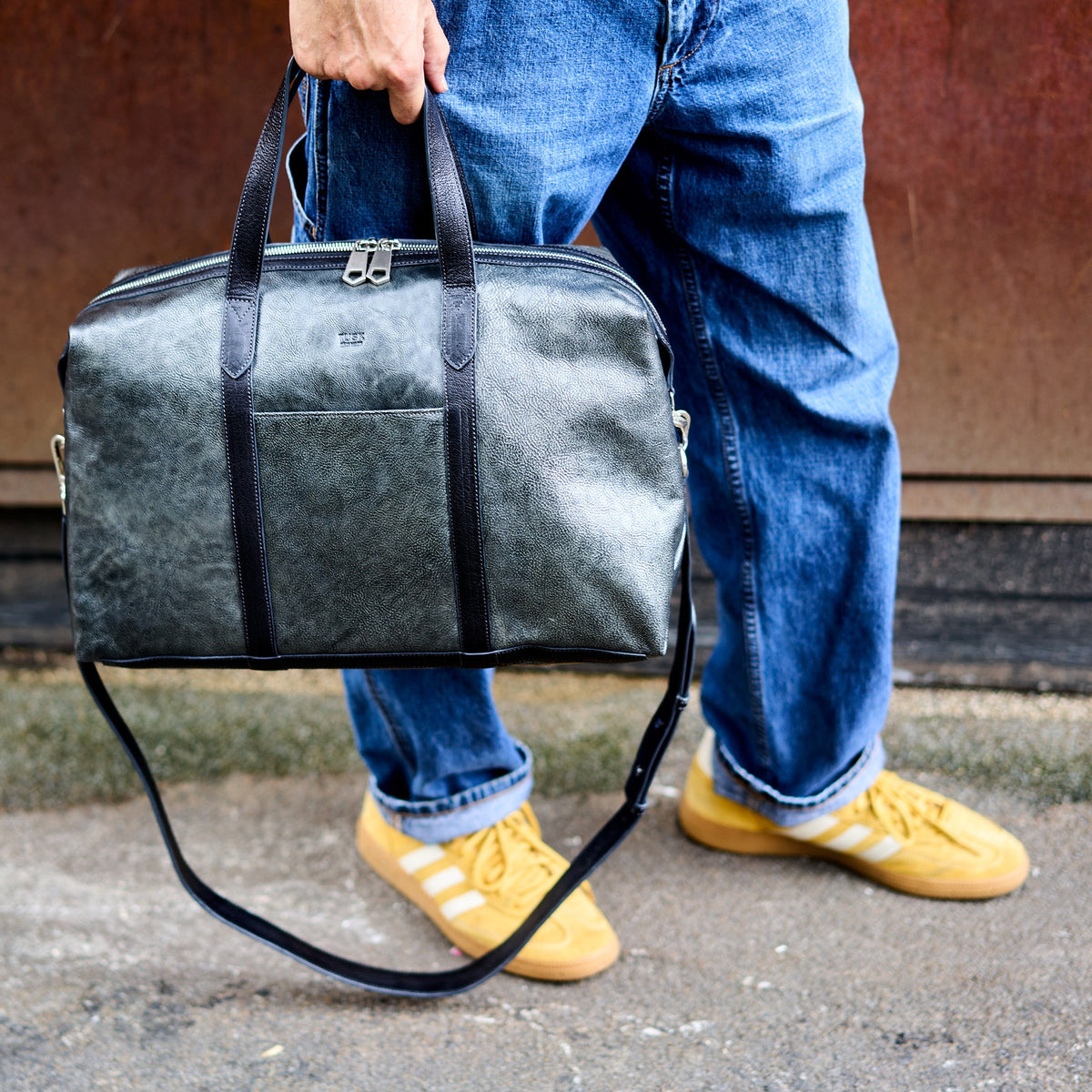 A person wearing blue jeans and yellow sneakers carries a dark gray textured leather duffel bag with black handles and a black shoulder strap - SILVER