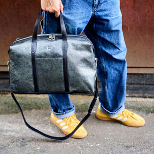A person wearing blue jeans and yellow sneakers carries a dark gray textured leather duffel bag with black handles and a black shoulder strap - SILVER