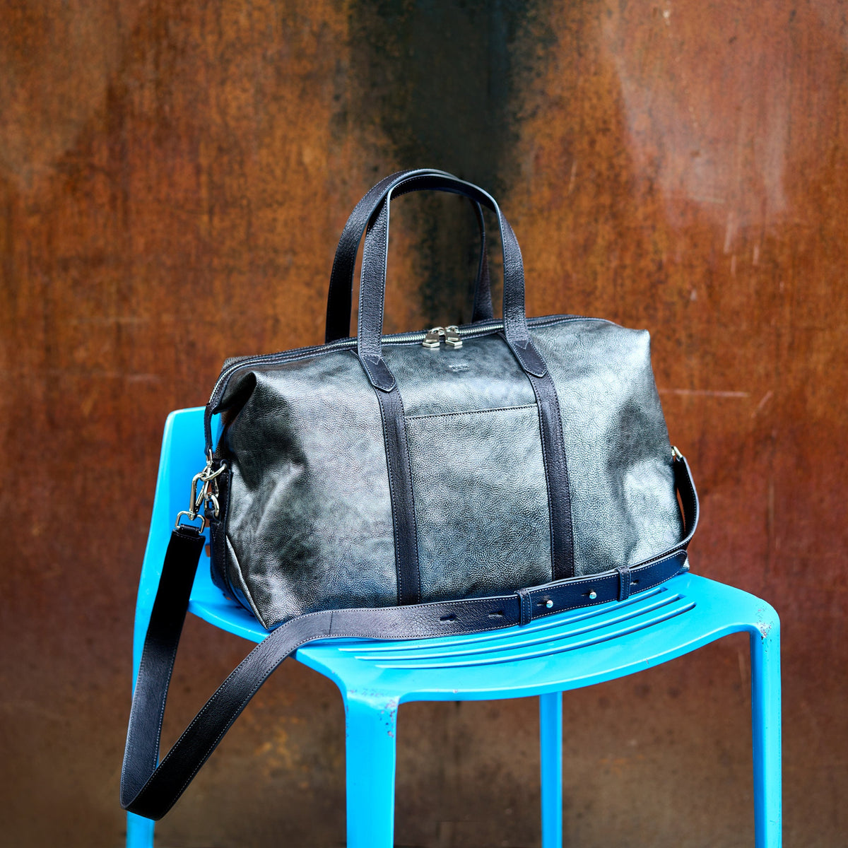A dark gray distressed leather duffel bag with black leather handles and trim, resting on a bright blue plastic chair against a textured rusted metal wall - SILVER