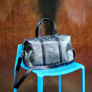 A dark gray distressed leather duffel bag with black leather handles and trim, resting on a bright blue plastic chair against a textured rusted metal wall - SILVER