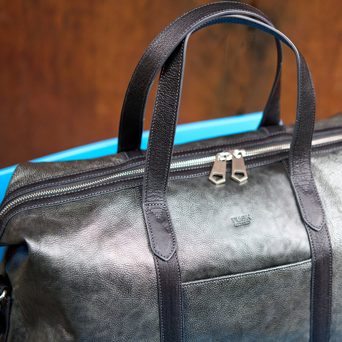 A close-up of a dark gray textured leather duffel bag with black handles and silver zipper pulls, resting on a bright blue surface against a warm brown background - SILVER