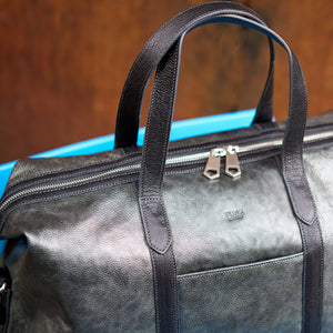 A close-up of a dark gray textured leather duffel bag with black handles and silver zipper pulls, resting on a bright blue surface against a warm brown background - SILVER