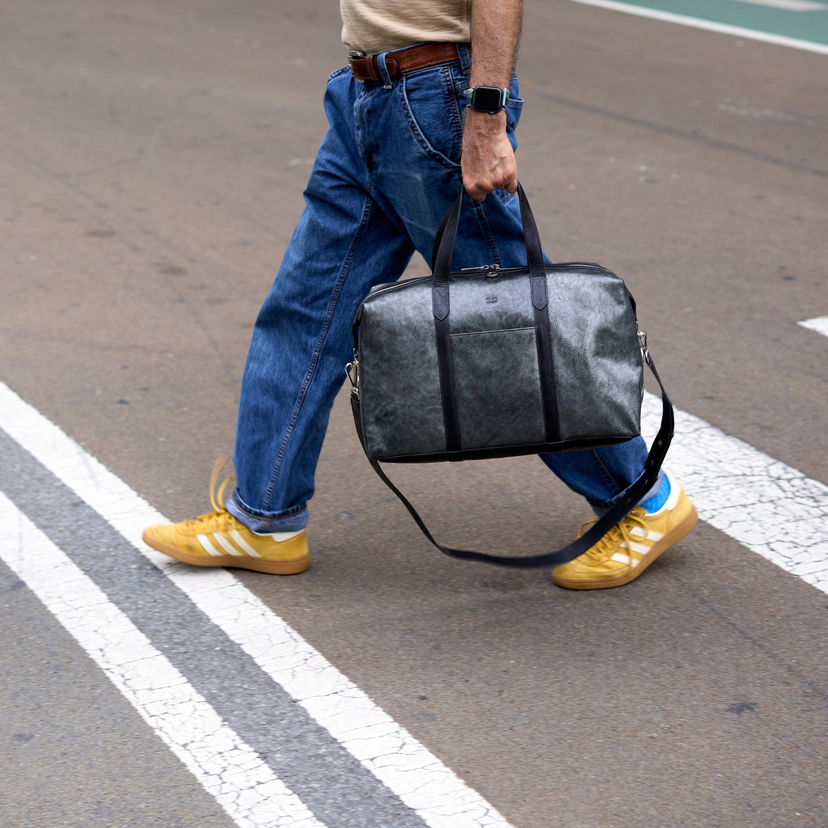 A person walking across a street crosswalk carrying a dark gray textured duffel bag with black straps. The person is wearing blue jeans, a beige t-shirt, and yellow sneakers - SILVER