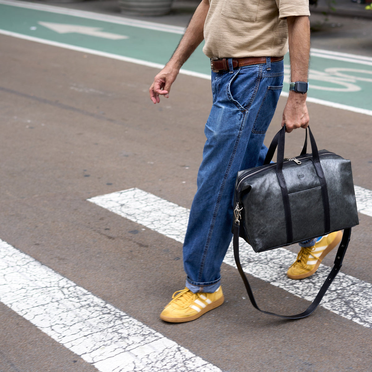 A man walking on a crosswalk carrying a dark gray distressed leather duffel bag. He is wearing blue jeans, a beige t-shirt, and yellow sneakers - SILVER