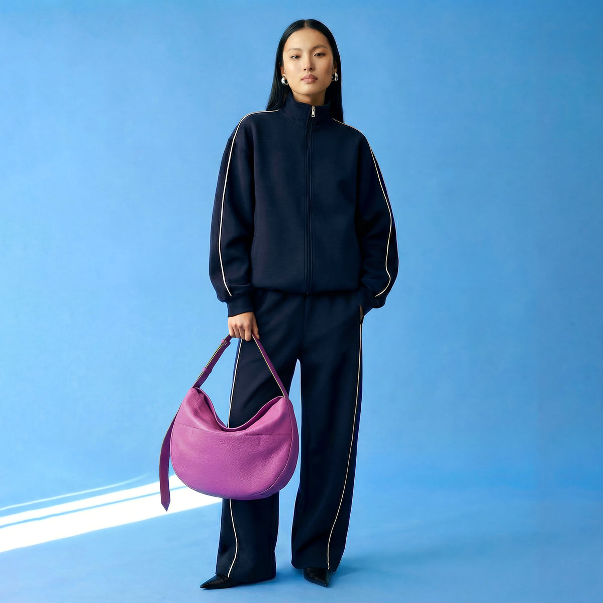 Person wearing a black outfit with white stripes, holding a purple handbag against a blue background - MAGENTA