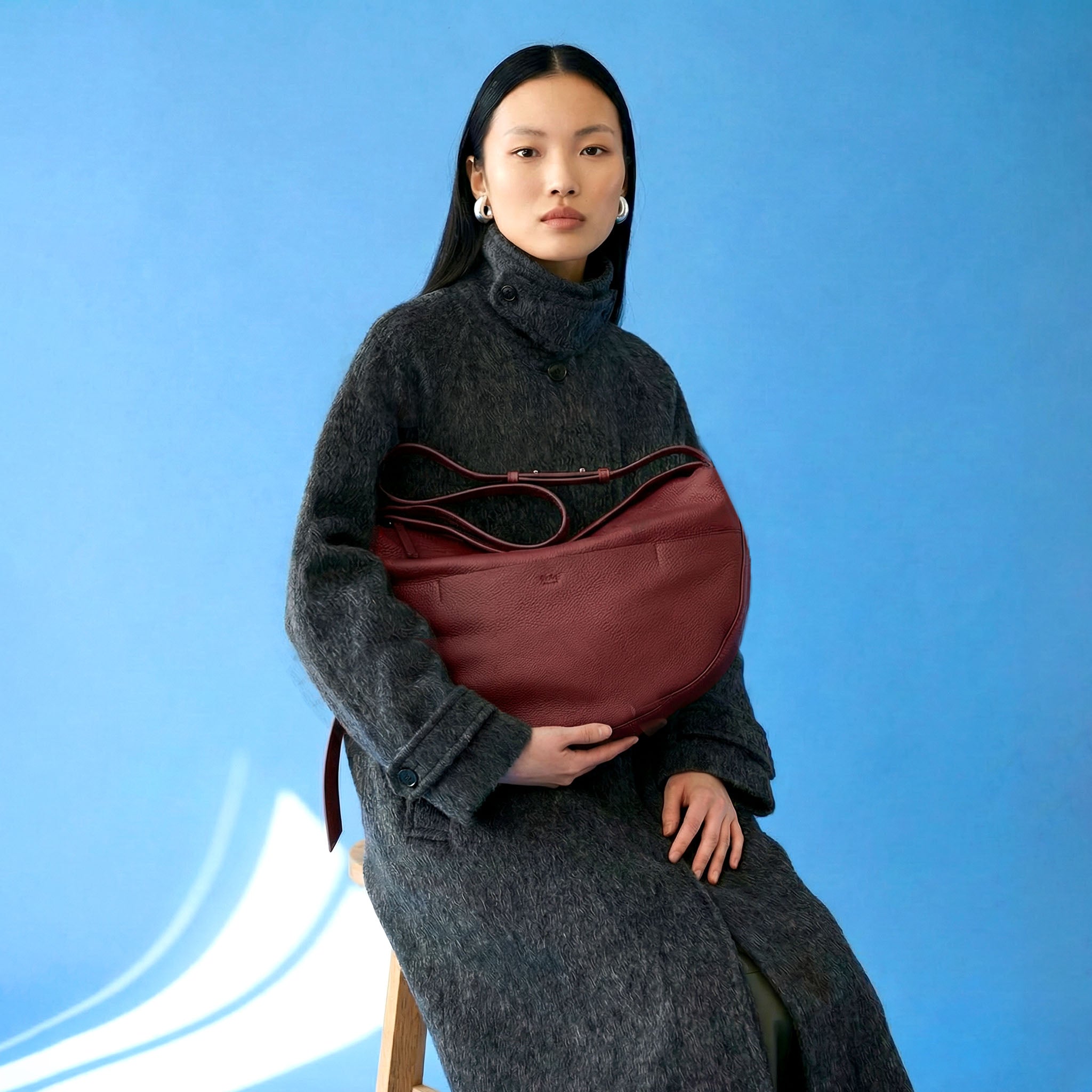 Woman holding a maroon handbag against a blue background - OXBLOOD