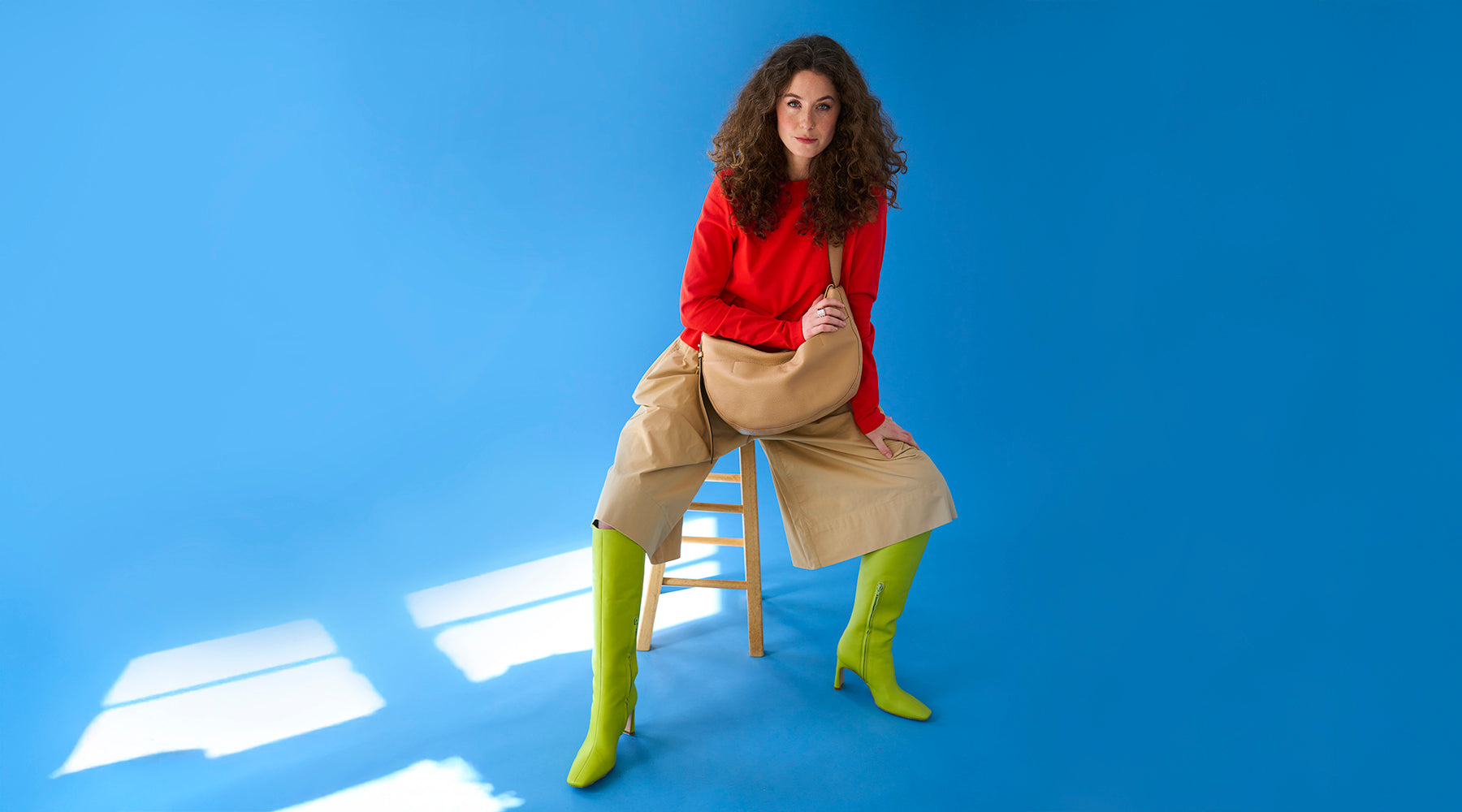 Woman wearing a red top, beige wide-leg pants, and neon green boots, sitting on a wooden stool with a tan Mira Large Hobo bag against a blue background