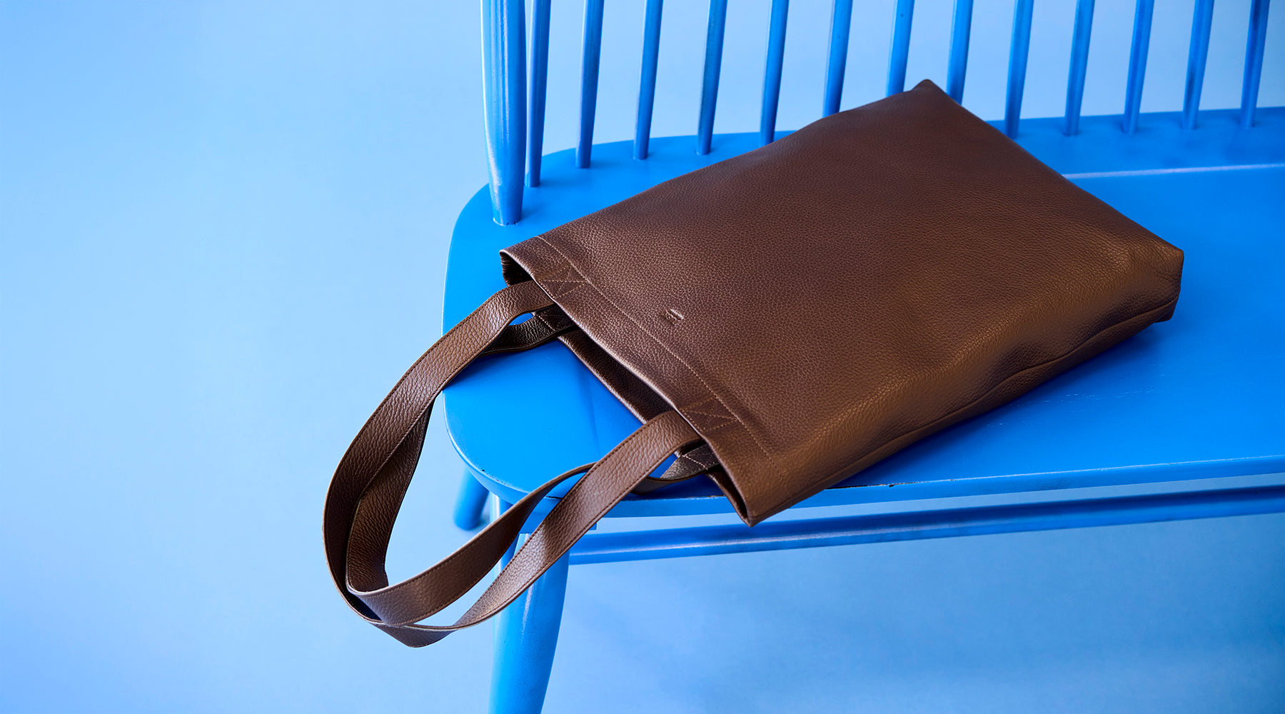 Brown leather bag with simple, minimalist design placed on a vibrant blue bench