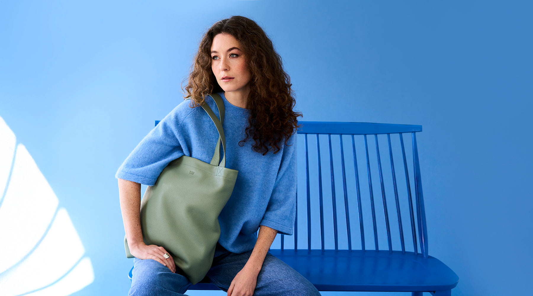 Woman sitting on a blue bench wearing a blue sweater and jeans, holding a soft green leather tote bag with the caption 'Our Best Selling Tote' displayed in the corner