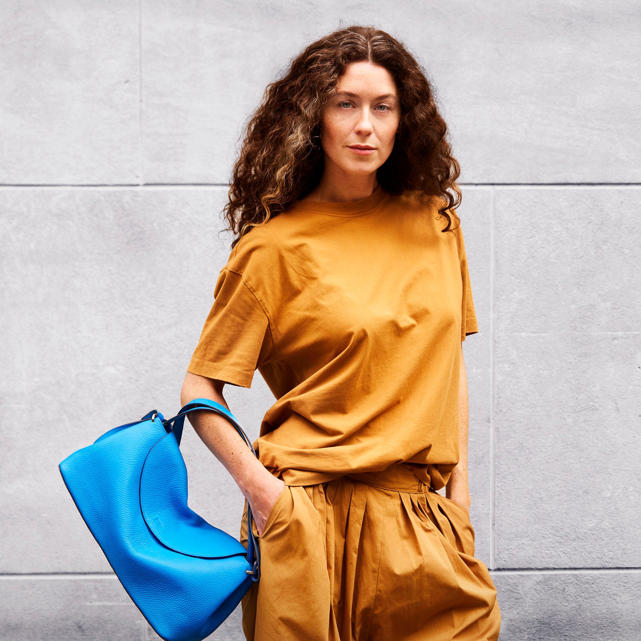 woman in mustard pant and shirt holding a blue italian leather bag on her arm on a neutral background - AZURE