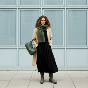 Woman wearing a beige coat, black dress, green scarf, and green rain boots standing against a light blue wall - FOREST