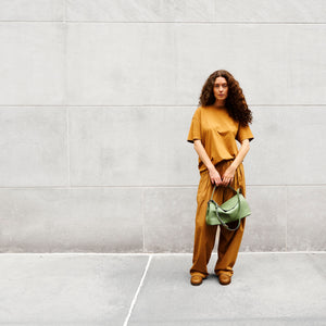 Woman in mustard yellow outfit holding a green handbag against a gray wall - SAGE