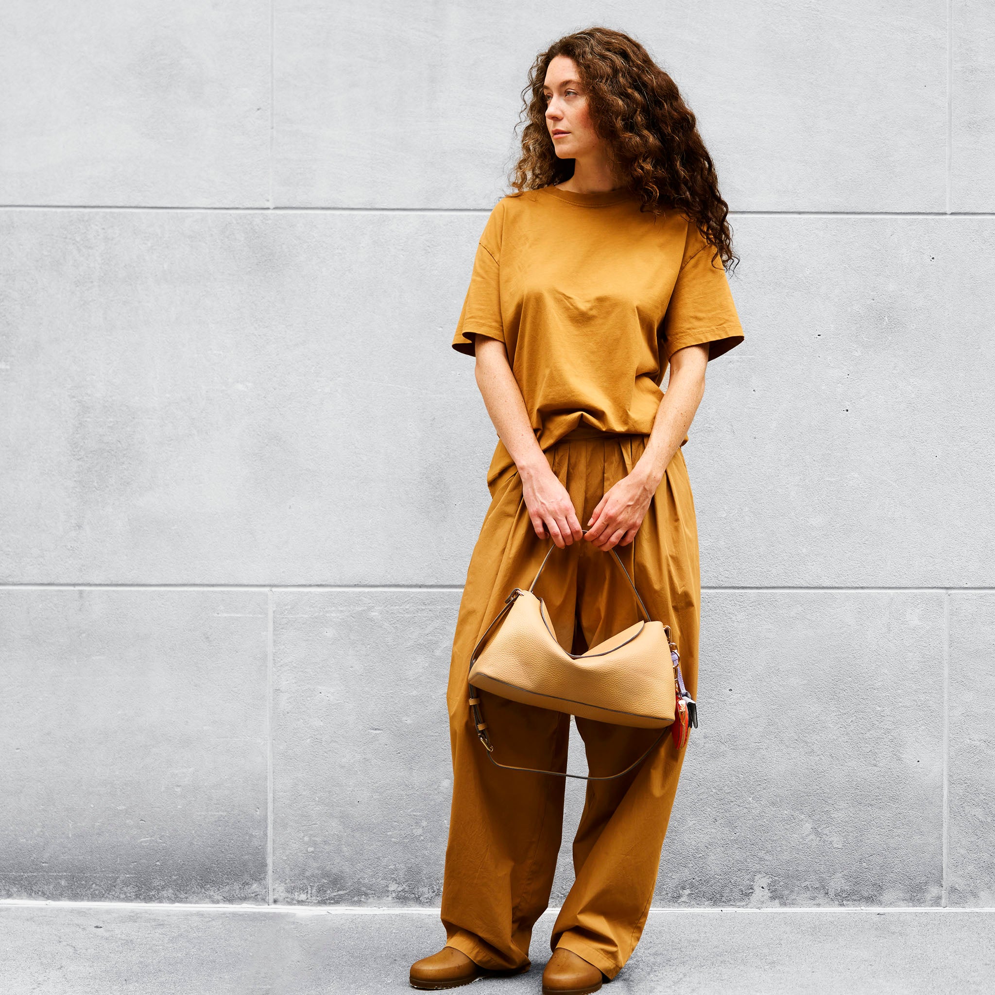 Woman in mustard yellow outfit holding a matching leather handbag against a gray wall - SAND