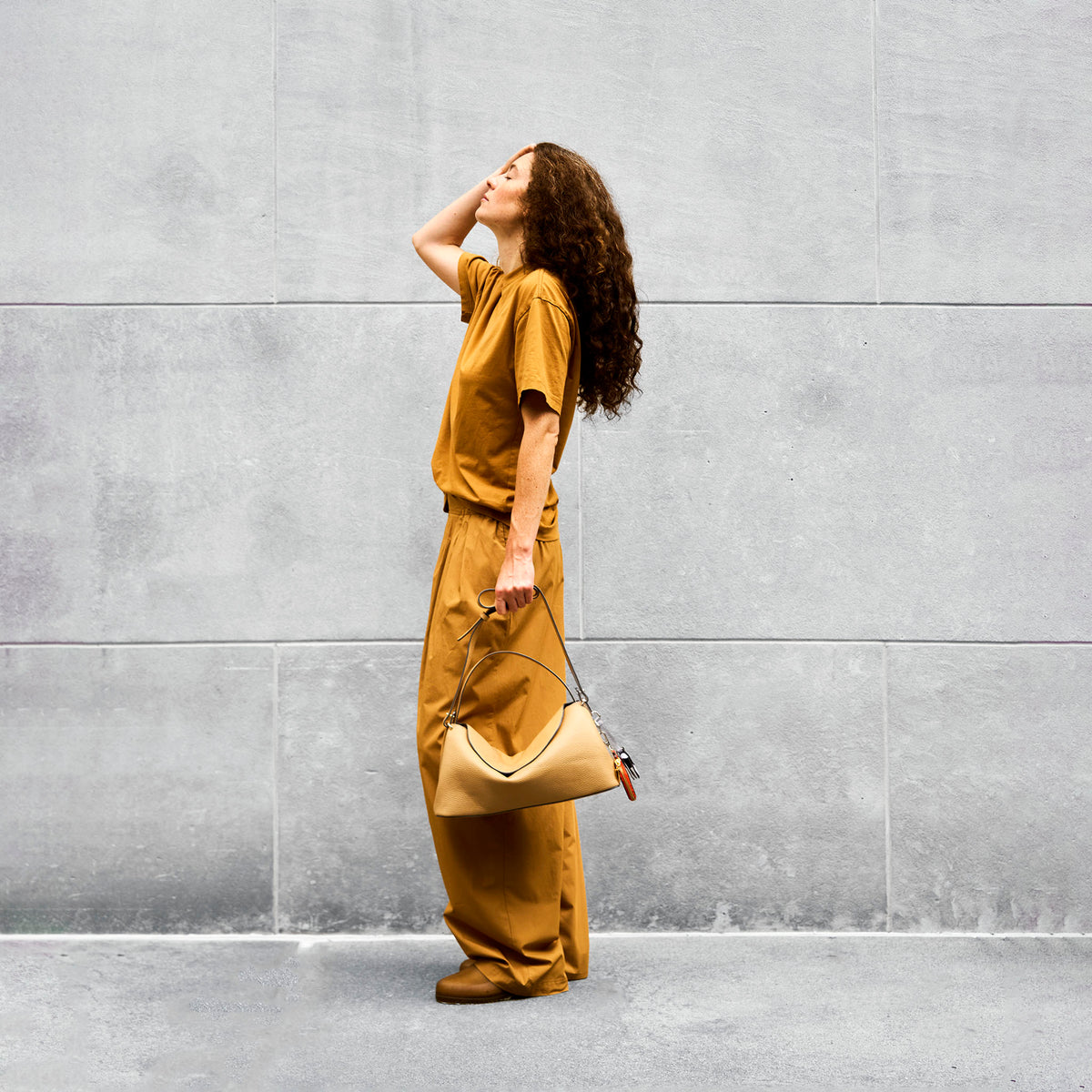 Woman in a mustard yellow outfit holding a matching leather handbag standing against a gray wall - SAND