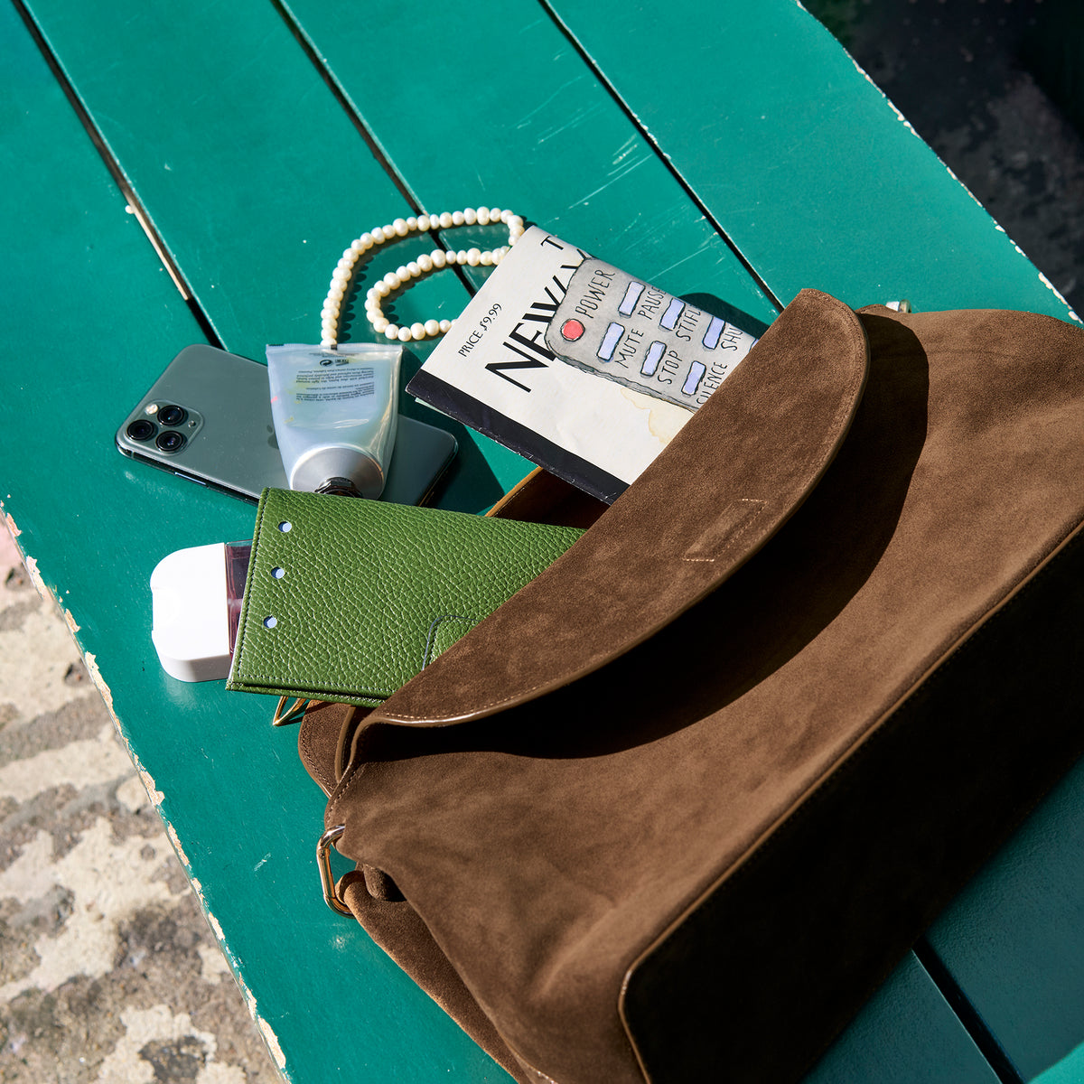 Brown leather bag with items including a phone, hand creme and green wallet on a green surface - BROWN