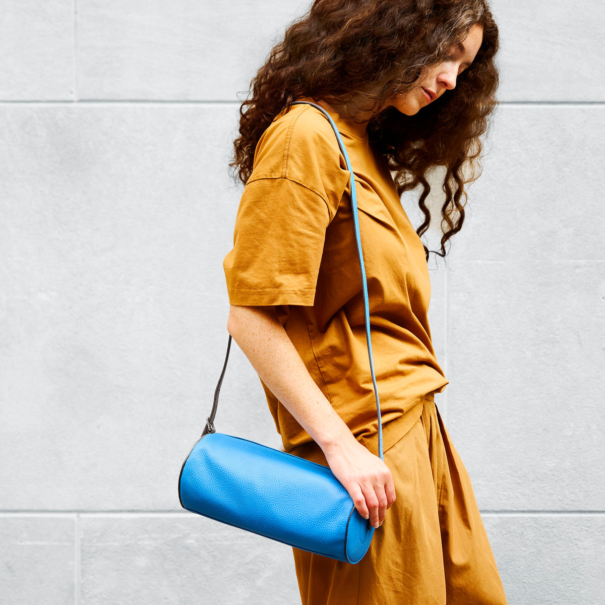 Woman carrying a blue leather handbag on her shoulder against a light gray wall - AZURE