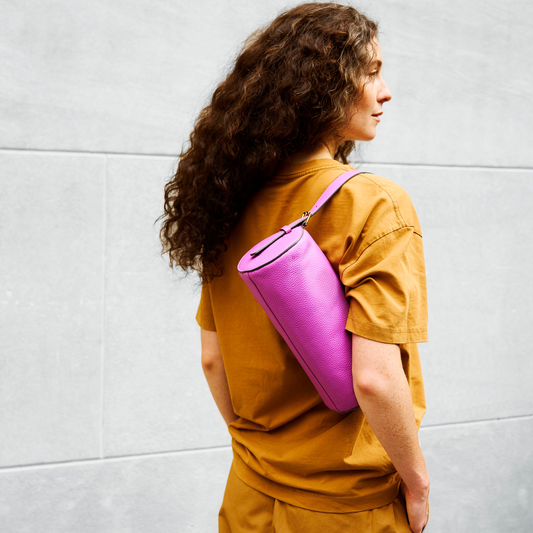 person wearing a mustard yellow shirt with a pink yoga mat over their shoulder against a light gray wall - MAGENTA