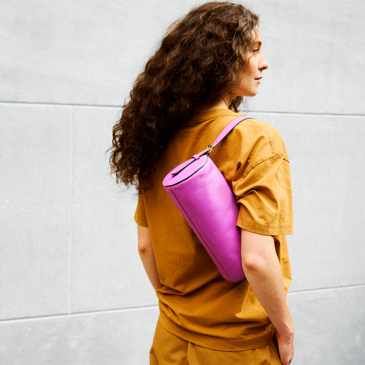 person wearing a mustard yellow shirt with a pink yoga mat over their shoulder against a light gray wall - MAGENTA