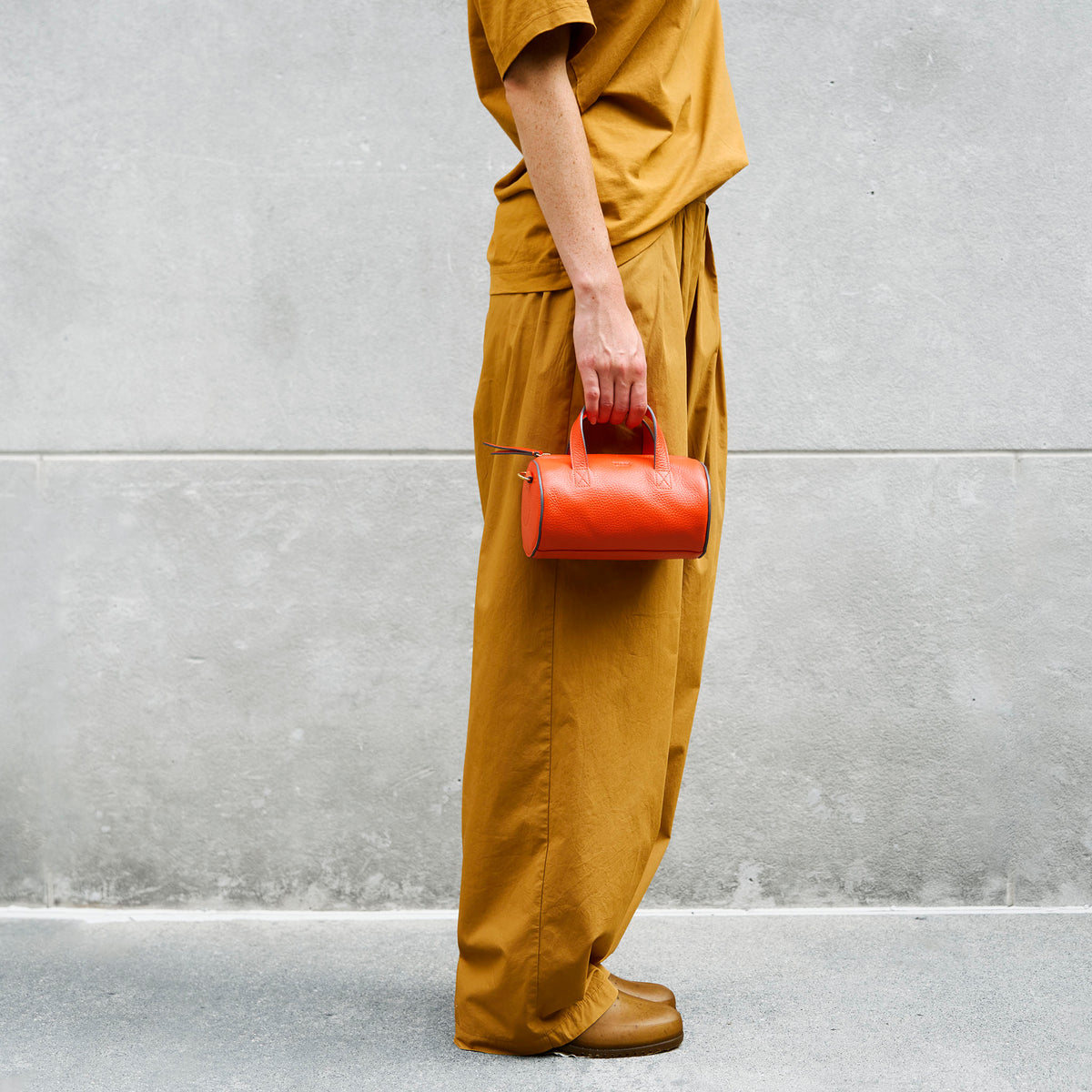 Person wearing mustard yellow outfit holding a orange leather handbag against a gray wall - ORANGE