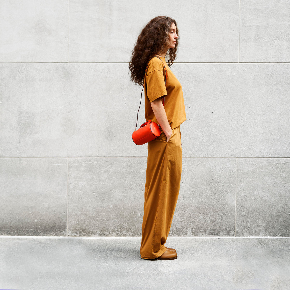 Woman in mustard yellow outfit holding a orange bag against a gray wall - ORANGE