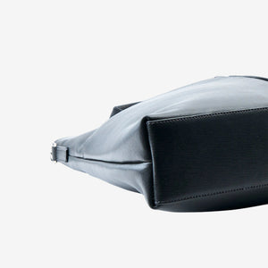A close-up side view of a grey leather handbag or satchel on a plain white background, showing the texture of the leather and a silver-toned metal zipper pull - CHARCOAL