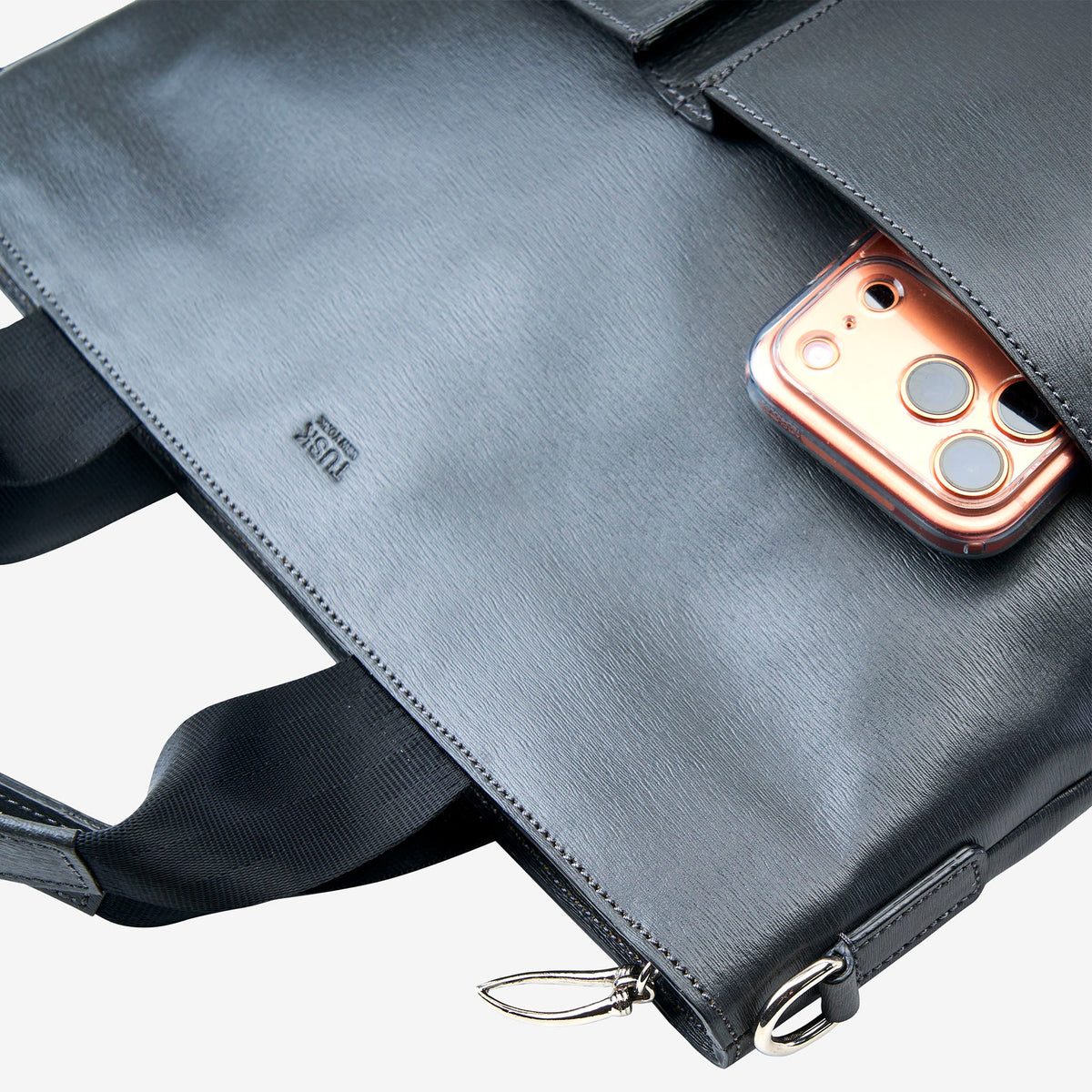 A close-up of a grey leather briefcase or laptop bag with a zippered pocket holding a rose gold smartphone. The brand name 'TUSK' is debossed on the leather - CHARCOAL