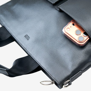 A close-up of a grey leather briefcase or laptop bag with a zippered pocket holding a rose gold smartphone. The brand name 'TUSK' is debossed on the leather - CHARCOAL