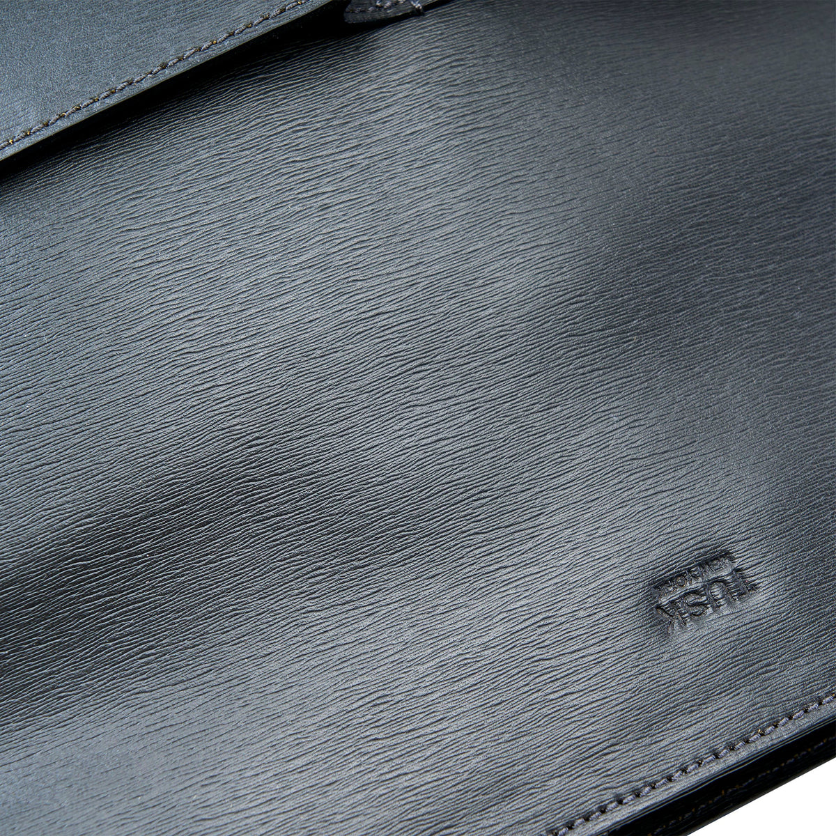 Close-up of a dark gray textured leather bag with visible grain and a small debossed logo near the lower right.
