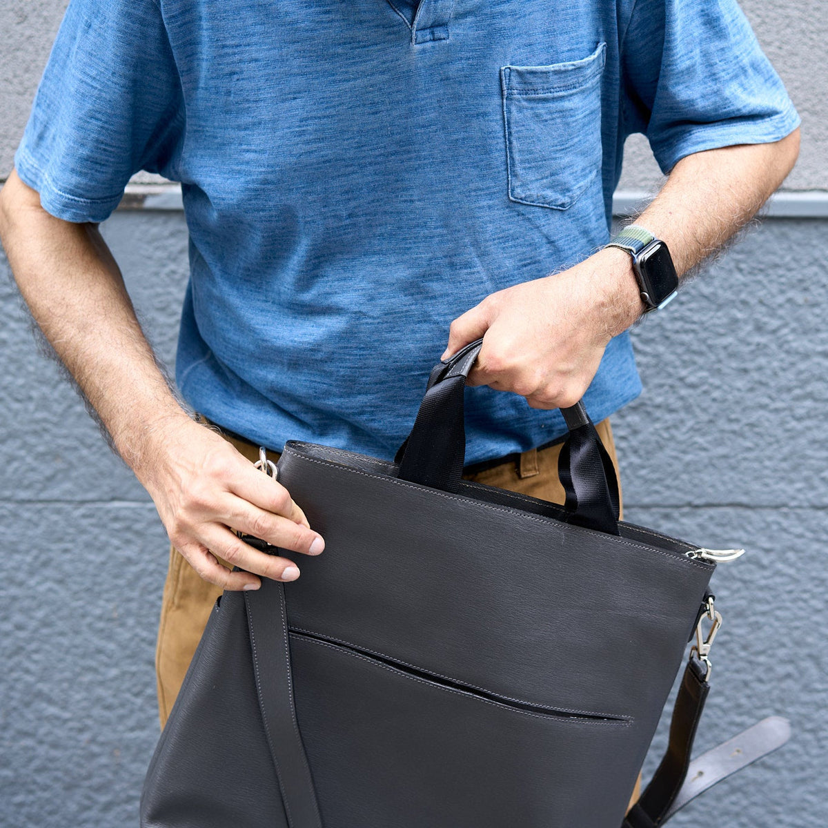Person holding a black bag against a gray wall - CHARCOAL