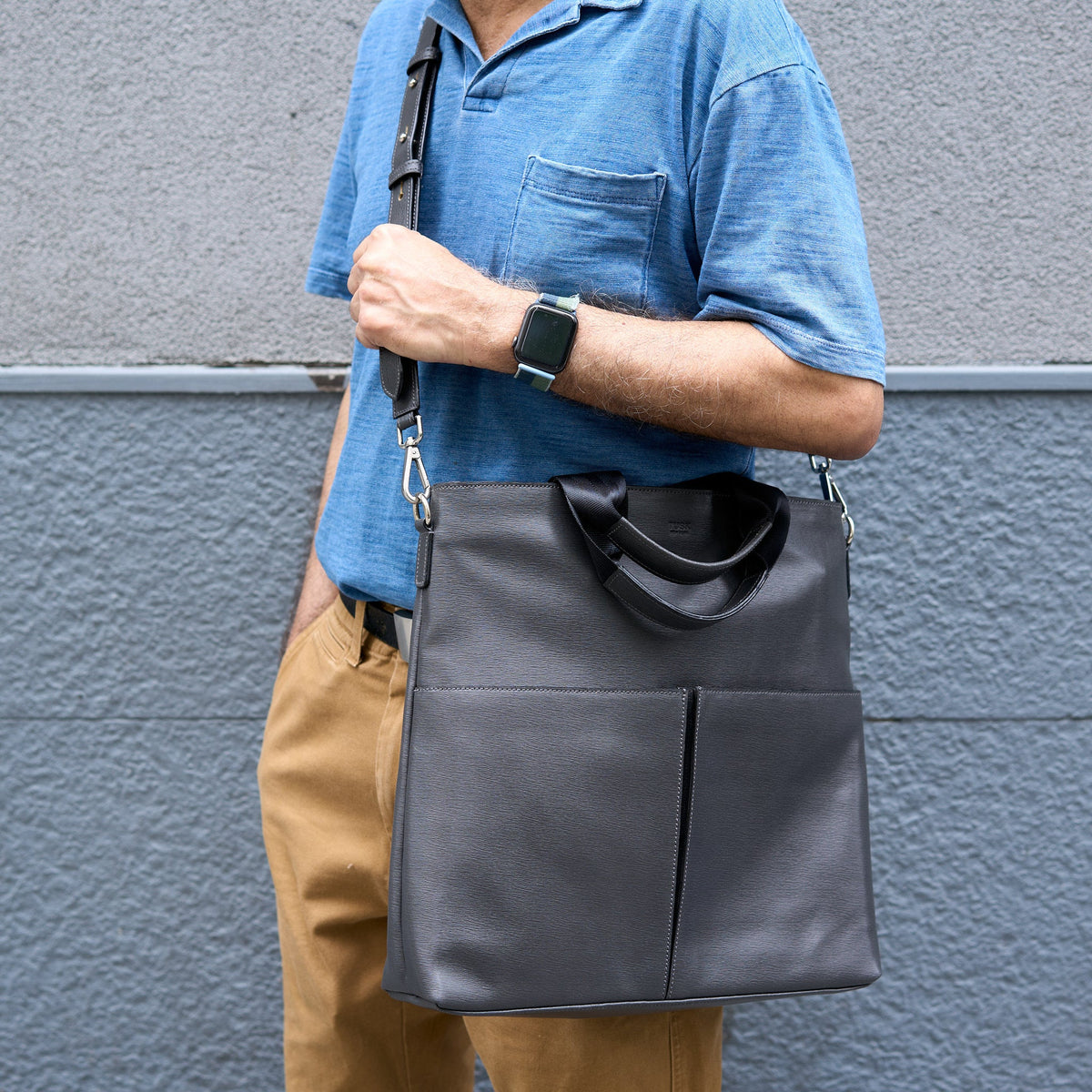 Person holding a dark gray leather bag against a gray wall - CHARCOAL