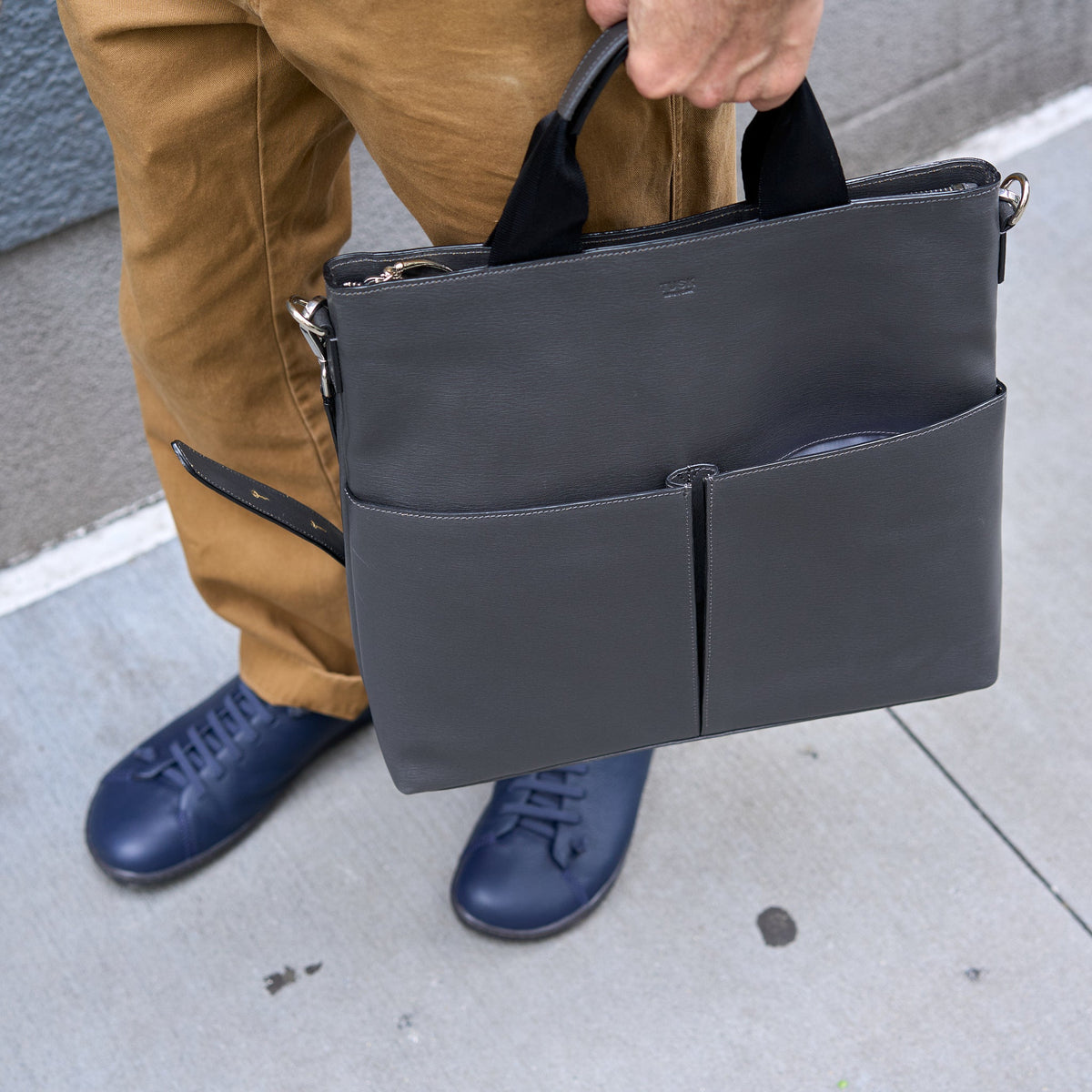 Person holding a dark gray leather bag on a sidewalk - CHARCOAL