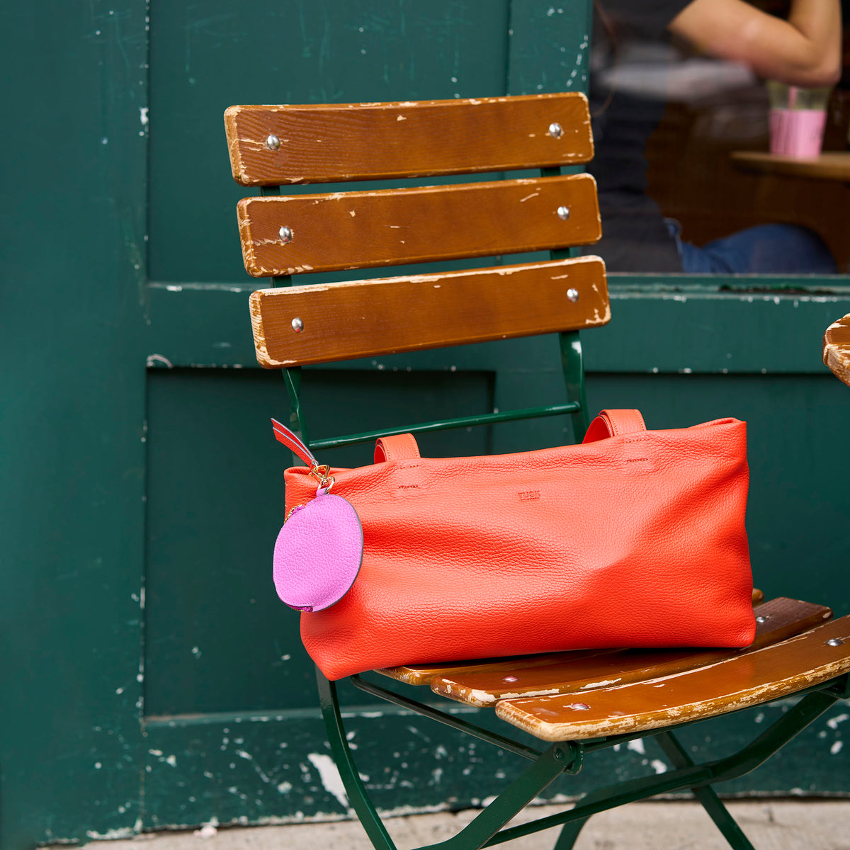 Orange handbag with a pink tag on a wooden chair against a green wall -ORANGE