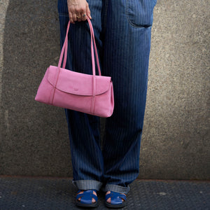 A person wearing blue pinstripe jeans and blue sandals holds a pink leather shoulder bag against a textured gray concrete wall - BLUSH