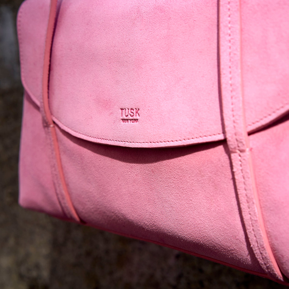 A close-up of a pink suede TUSK small shoulder bag with a flap closure and visible 'TUSK NEW YORK' branding, set against a dark textured background -  BLUSH