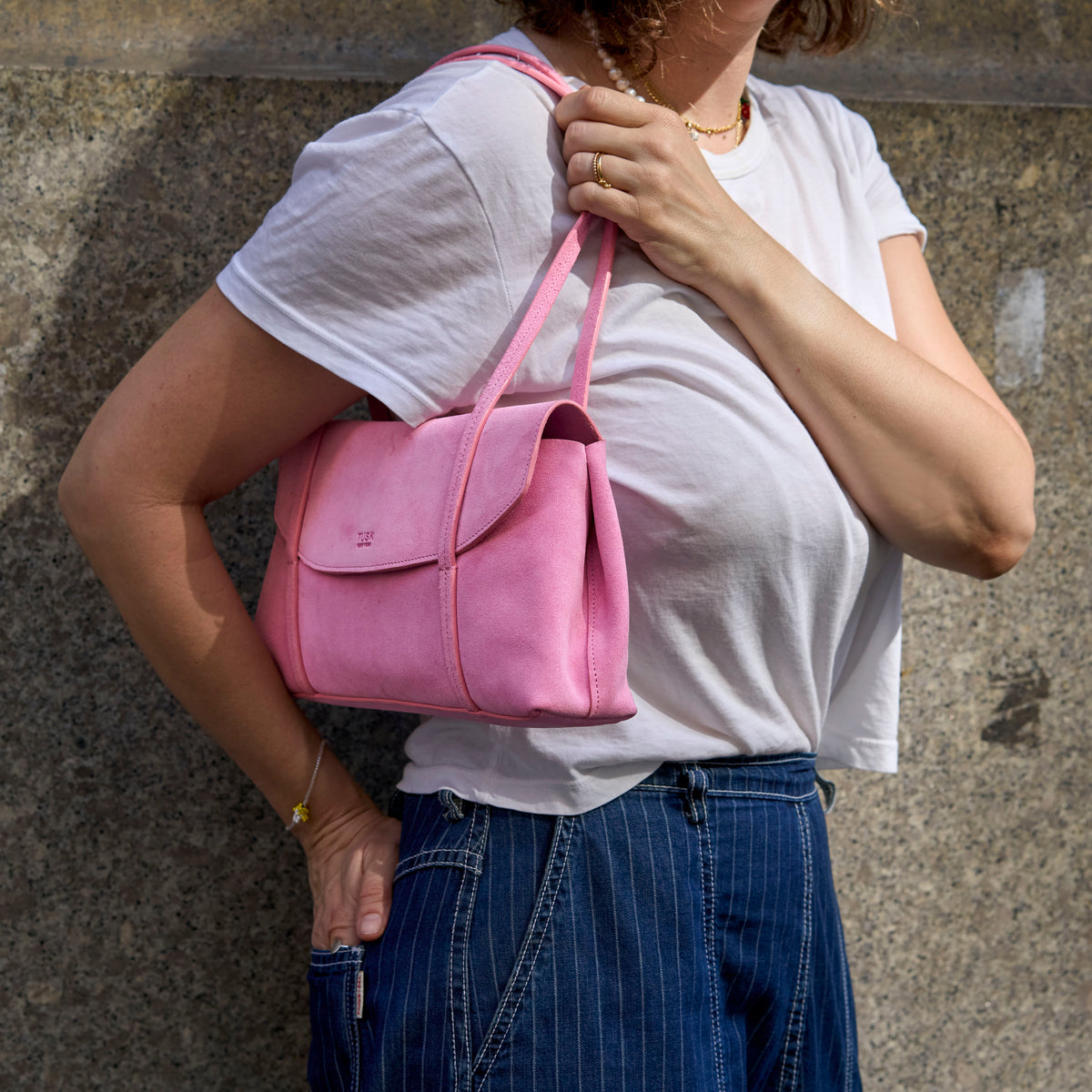 A woman wearing a white t-shirt and blue pinstripe pants carries a pink suede shoulder bag against a textured gray concrete wall -  BLUSH