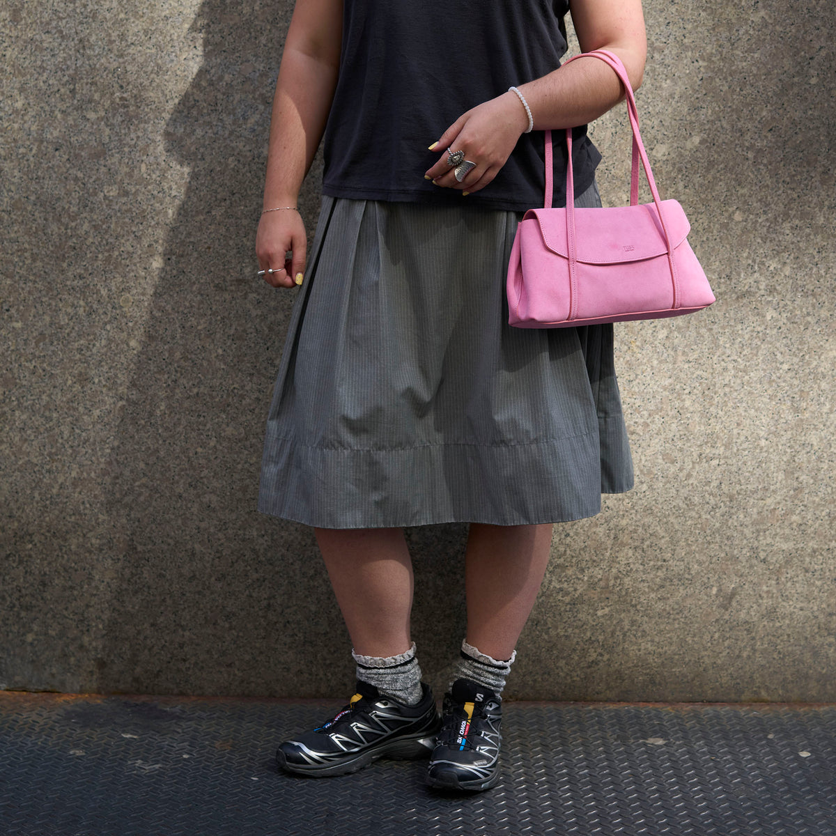 A person wearing a dark gray pleated midi skirt, black t-shirt, gray socks, and black Salomon trail running shoes, carrying a pink leather shoulder bag against a textured concrete wall -  BLUSH